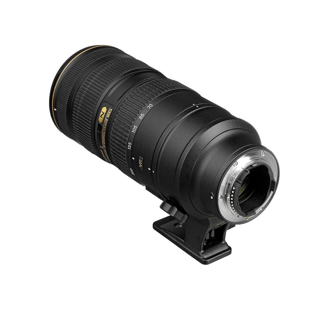 af-s_nikkor_70-200mm_f2_8g_ed_vrii_0001 Nikon AF-S Nikkor 70-200mm f/2.8G ED VR II -teleobjektiivi, jossa on zoomaus- ja tarkennusrengas sekä jalustakiinnityskaulus, on kuvattu valkoisella taustalla kulmassa, ja siinä on näkyviä kultaisia korostuksia ja objektiivin merkintöjä.