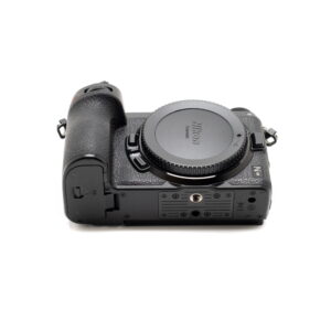 Käytetty musta Nikon Z6 II (Kunto K5, Takuuta 22kk, Shuttercount 1100) -kamerarunko on kuvattu selällään makaavana, jolloin objektiivin kiinnityssuoja, kuvioitu kahva sekä eri portit ja pohjan ruuvikiinnikkeet tulevat esiin.