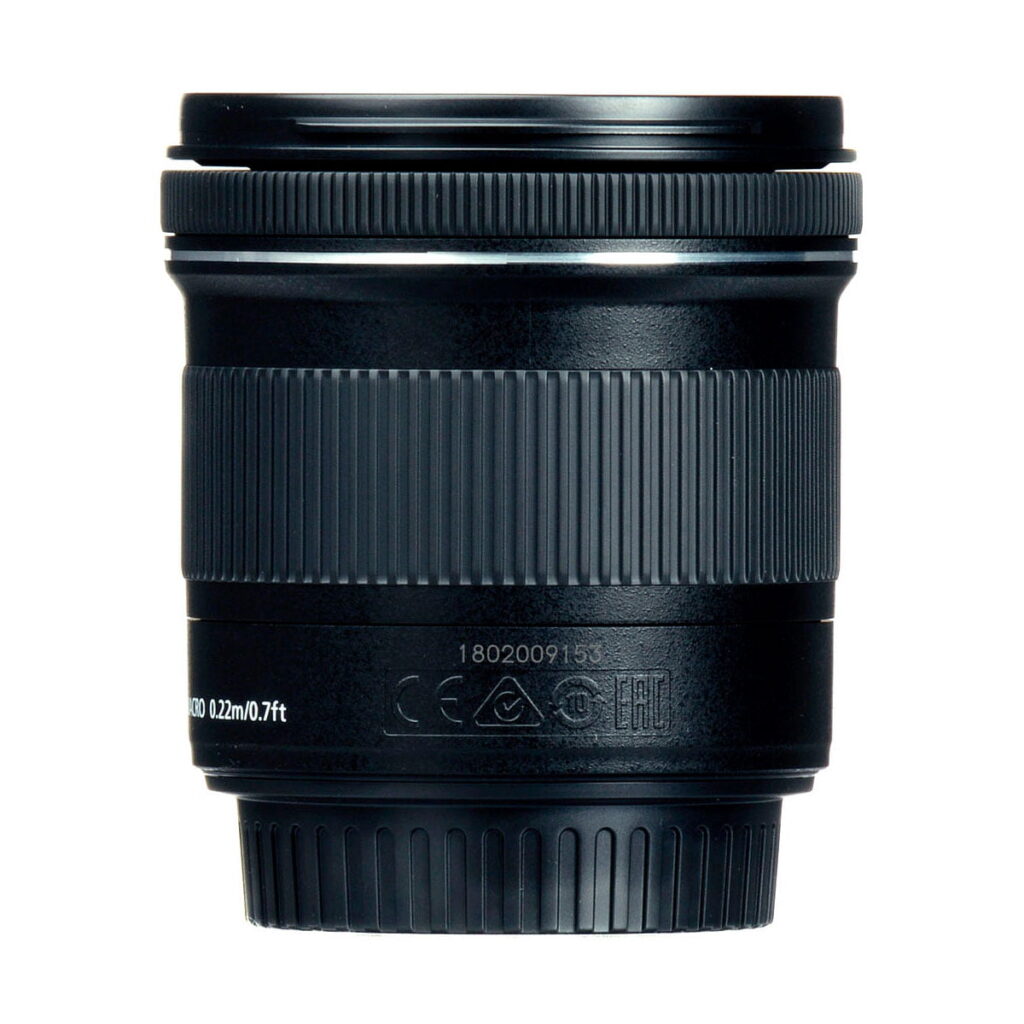 Canon_EF-S_10-18mm_f_4-5_6_IS_STM_003 Lähikuva Canon EF-S 10-18mm f/4.5-5.6 IS STM -objektiivista näyttää sen mustat, kuvioidut tarkennusrenkaat ja zoomausrenkaat, joissa on valkoisia merkintöjä ja symboleja lähellä pohjaa, kaikki valkoista taustaa vasten.