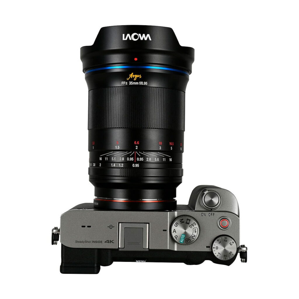 Laowa_Argus_35mm_f_0_95_FF-Sony-E_007 Ylhäältä katsottuna hopeinen digitaalikamera, jossa on säätöpyörät, painikkeet ja kuvioitu oikea kahva, johon on kiinnitetty suuri Laowa Argus 35mm f/0.95 FF - Sony-E -objektiivi.