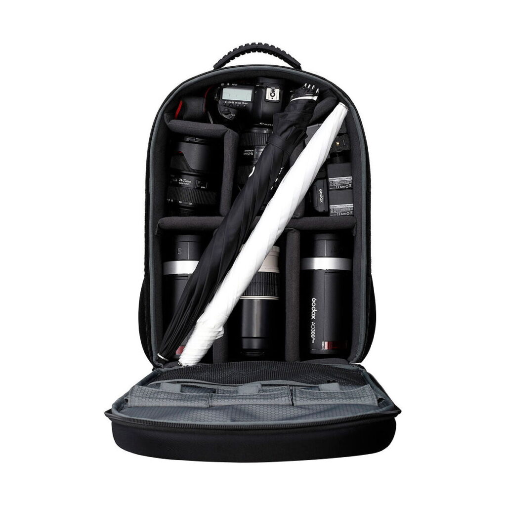 Godox_AD300_Outdoor_2-FLASH_Kit_001 Avoin kamerareppu, jossa on järjestetyt lokerot, joihin mahtuu useita kameran objektiiveja, kameran runko, akut, taitettu valokuvaussateenvarjo ja Godox AD300 Pro Outdoor 2-Flash Kit.