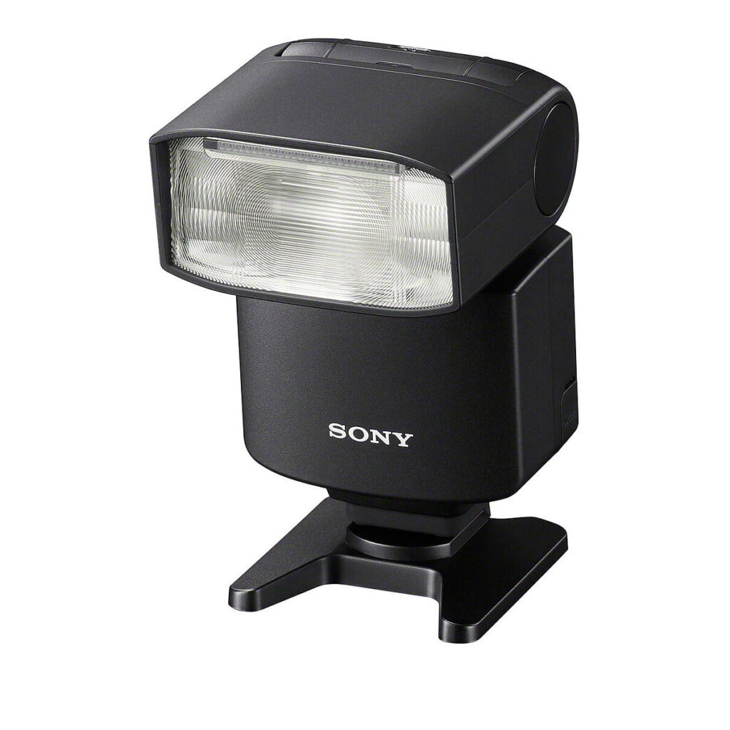 Sony_HVL-F46RM_007 Musta Sony HVL-F46RM -kameran ulkoinen salama, jossa on suorakulmainen pää ja leveä diffuusori ja joka on asennettu pieneen muovijalustaan, on esillä valkoista taustaa vasten.