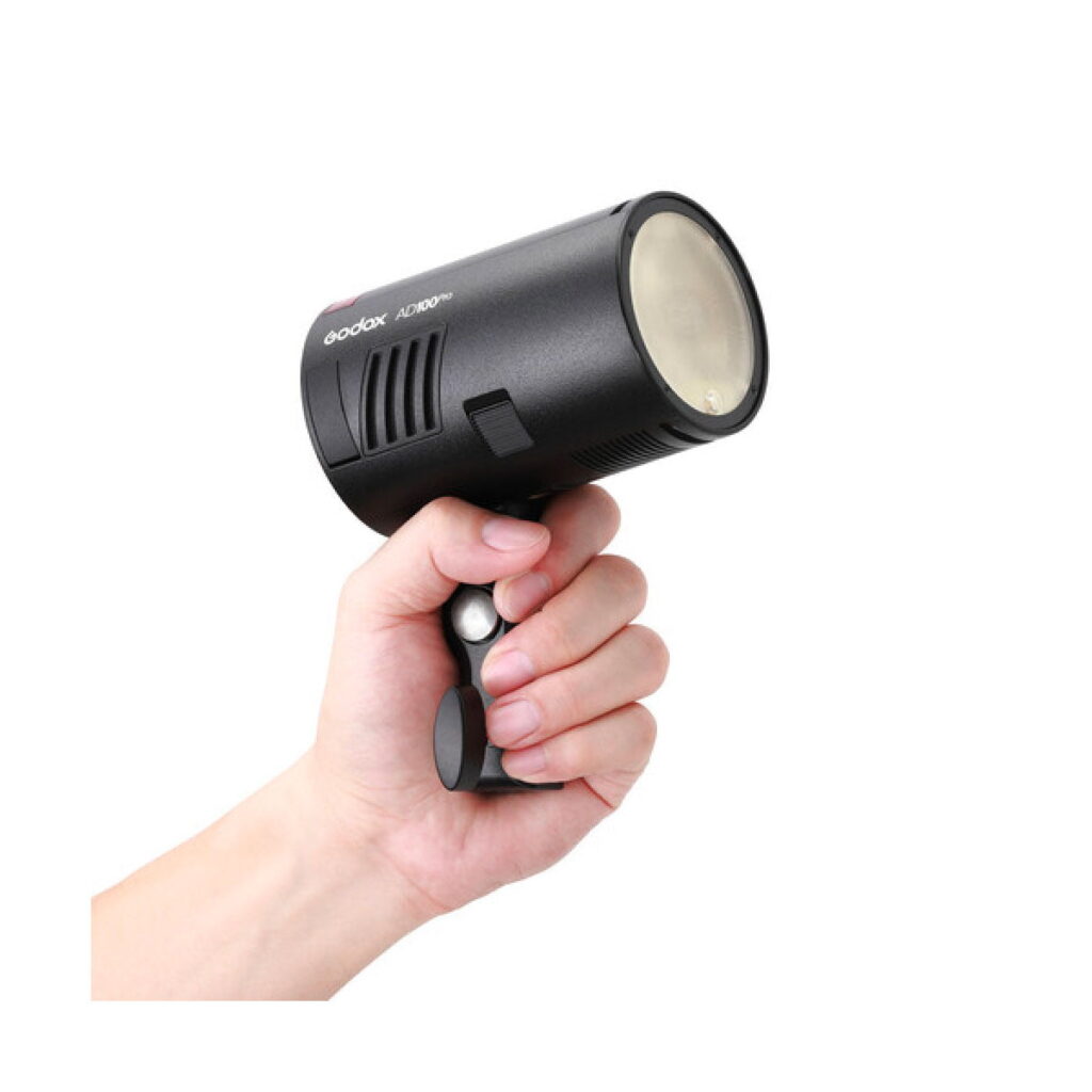 godox_ad100_pro_ttl_02 Käsi pitelee Godox AD100 Pro TTL -akkusalaman kannettavaa salamaa, jossa on lieriömäinen runko ja kahva, valkoista taustaa vasten.