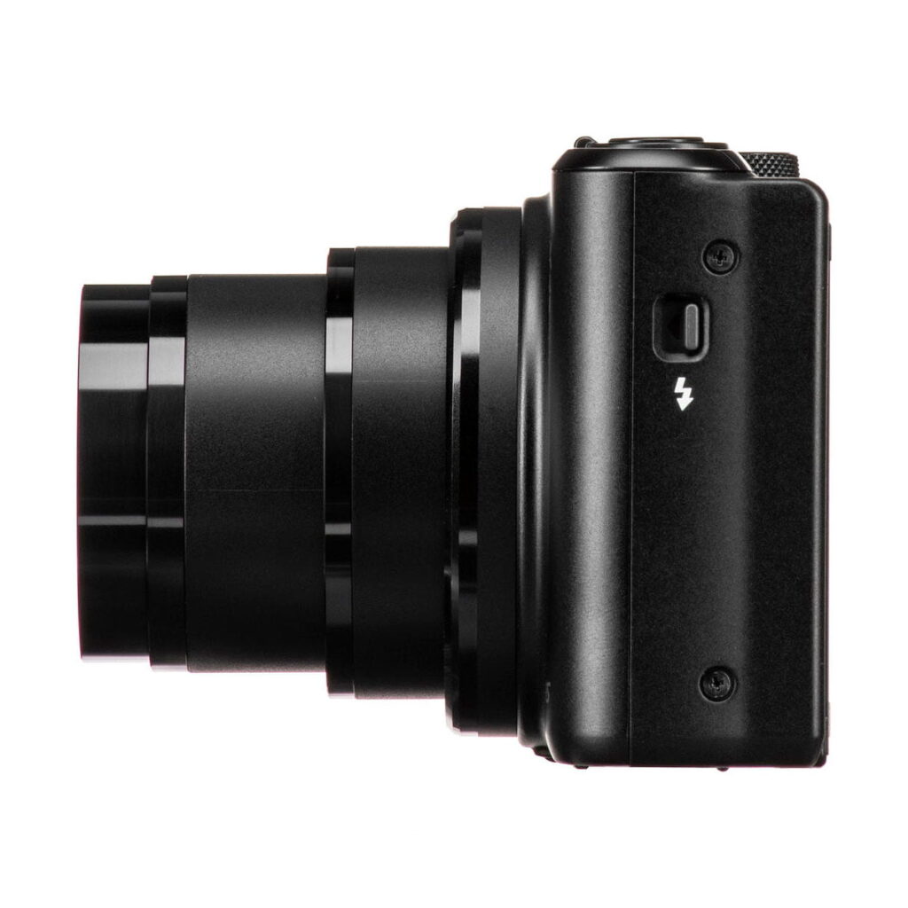 Mustan värinen Canon PowerShot SX740 HS -kompaktikamera, jonka objektiivi on ulosvedetty, kuvassa sivuttain valkoista taustaa vasten. Rungossa näkyy salamasymboli pienen painikkeen yläpuolella.