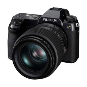 Fujifilm_GFX_100S_011 Musta Fujifilm GFX 100S -järjestelmäkamera, jossa on suuri irrotettava objektiivi, useita ohjauspainikkeita ja valintakiekkoja, näkyy vinossa valkoista taustaa vasten.