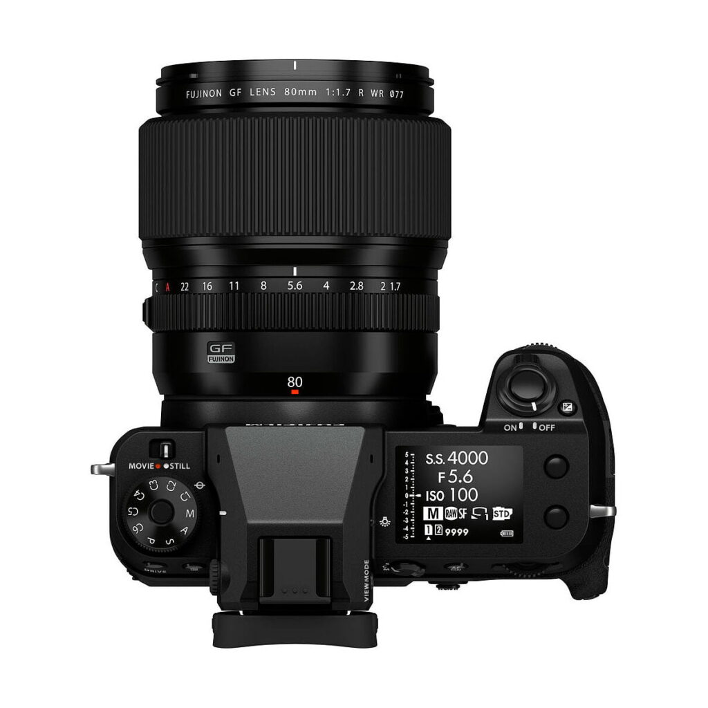 Fujifilm_GFX_100S_013 Ylhäältä katsottuna musta Fujifilm GFX 100S -järjestelmäkamera, jossa on suuri 80mm objektiivi, jossa näkyy säätöpyörät, painikkeet, elektroninen etsin ja näyttö, joka näyttää kuvausasetukset.