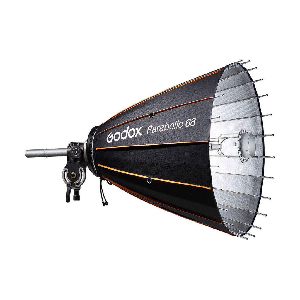 Godox_Parabolic_68_Reflector_Kit_001 Godox Parabolic 68 -heijastinsarja, musta oransseilla korostuksilla, on asennettu kiinnikkeeseen, jonka sivusta ulottuu hopeinen tanko ja joka näkyy valkoista taustaa vasten.