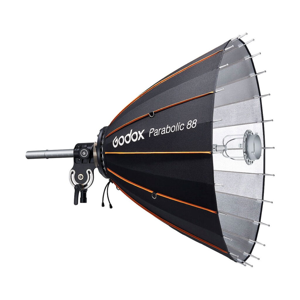 Godox_Parabolic_88_Reflector_Kit_002 Sivunäkymä Godox Parabolic 88 -heijastinsarjasta, jossa on musta ja hopeinen ulkokuori oransseilla reunuksilla, paljaat tangot ja joka on asennettu studiovalaisimeen.