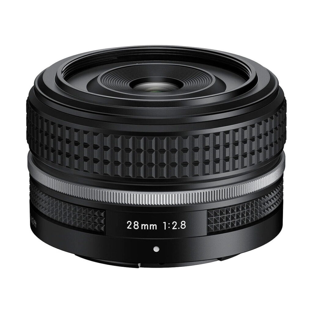 Nikon_Nikkor_Z_28mm_f_2_8_001 Nikon Nikkor Z 28mm f/2.8 SE -objektiivi on musta kameraobjektiivi, jossa on kuvioitu tarkennusrengas ja hopeinen korostus valkoista taustaa vasten.