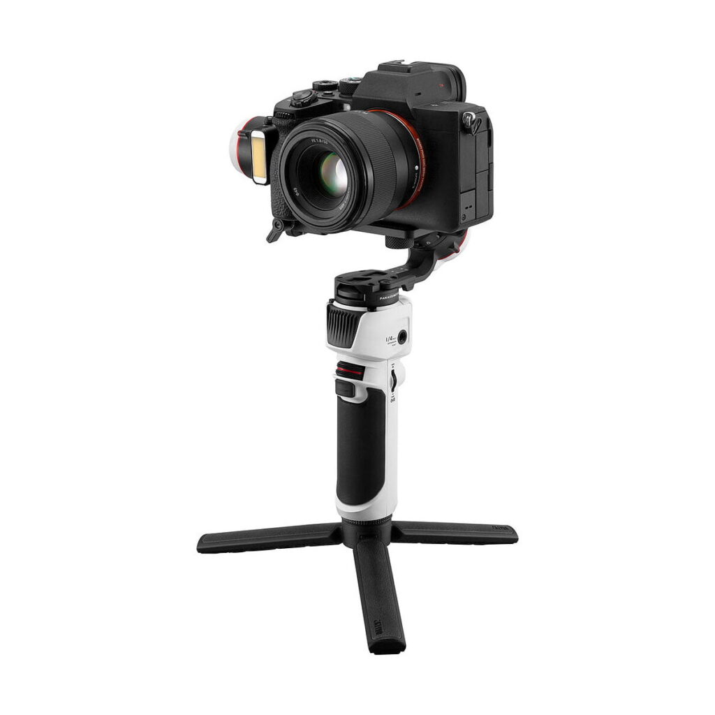 Zhiyun-Tech_Crane_M3_005 Zhiyun Crane M3 - Gimbaali on valkoinen käsikäyttöinen gimbal-stabilisaattori, jossa on musta kahva ja kokoontaitettavat jalustan jalat, kuvassa digitaalinen kamera asennettuna siihen valkoista taustaa vasten.