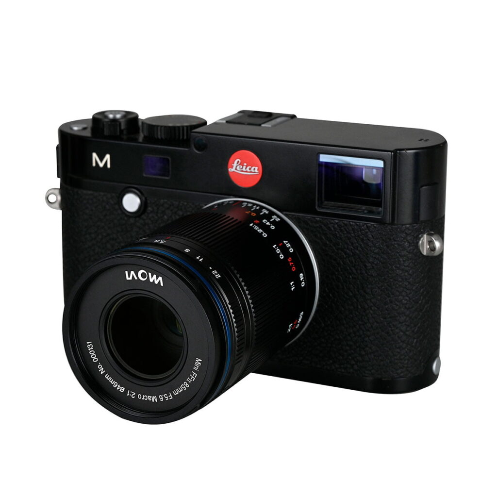 Laowa_85mm_f_5_6_2X_Ultra_Macro_APO_002 Musta Leica M -kamera, johon on kiinnitetty Laowa 85mm f/5.6 2X Ultra-Macro APO - Sony FE -objektiivi, kuvattuna kameran ja objektiivin etu- ja sivupuolelta valkoista taustaa vasten.