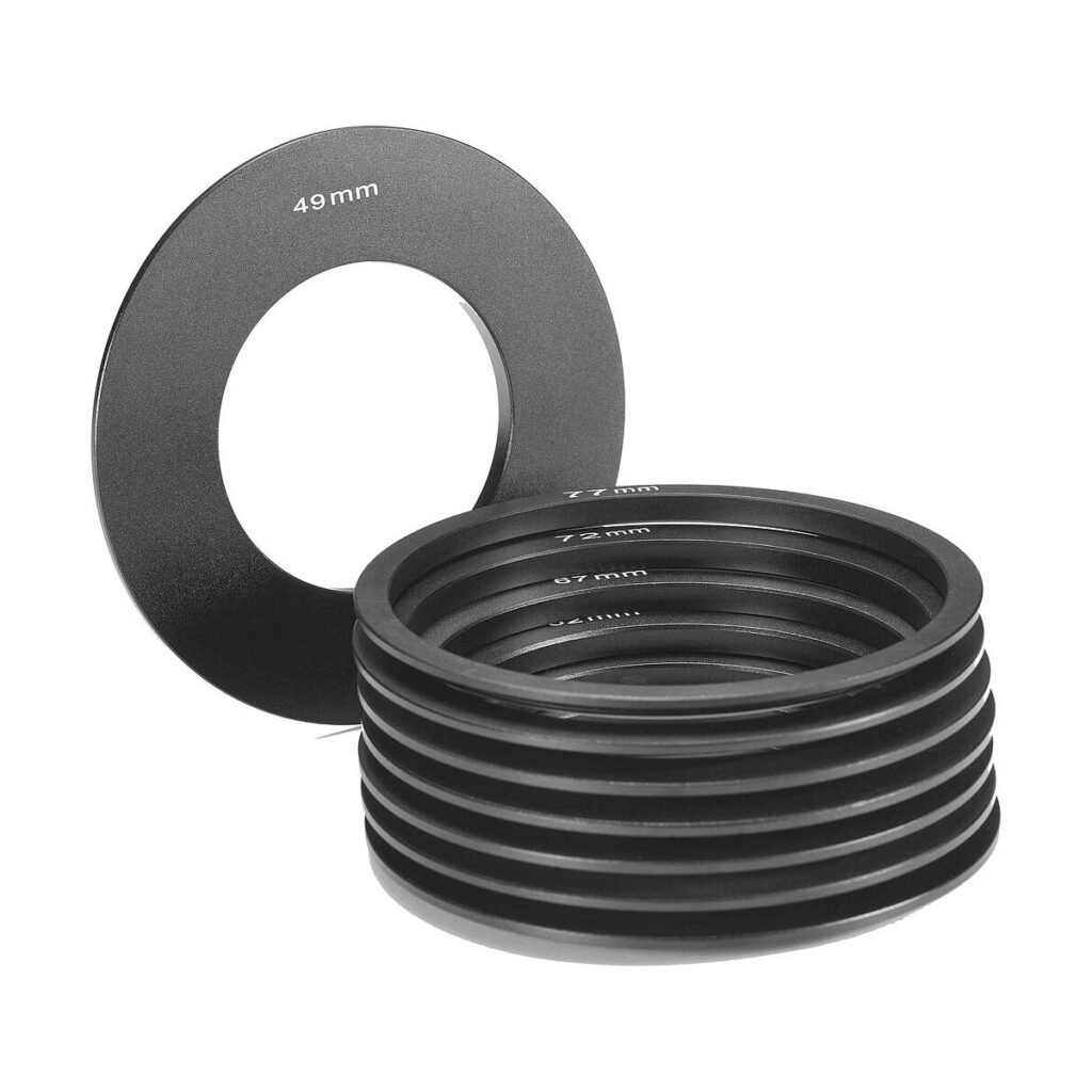 Godox_MF12_Set_of_Adapter_Rings_003 Joukko erikokoisia mustia metallisia Godox MF12 -sovitinrenkaita, joissa jokaisessa on merkintä 49 mm:n mittaisista mitoista, on pinottu päällekkäin, ja yksi rengas seisoo pystyssä Godox AD300 Pro -valaisimen vieressä.