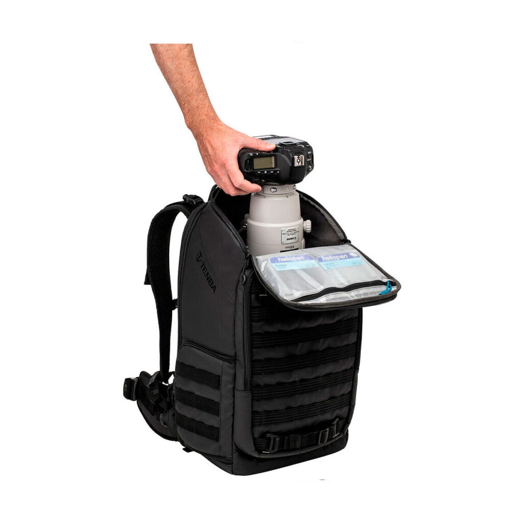 Tenba_Axis_20L_Tactical_Backpack_006 Henkilö asettaa DSLR-kameran, jossa on suuri teleobjektiivi, avoimeen mustaan Tenba Axis 20L Tactical Backpack -reppuun, jossa on järjestäytyneet sisätilat ja vetoketjulliset verkkotaskut.