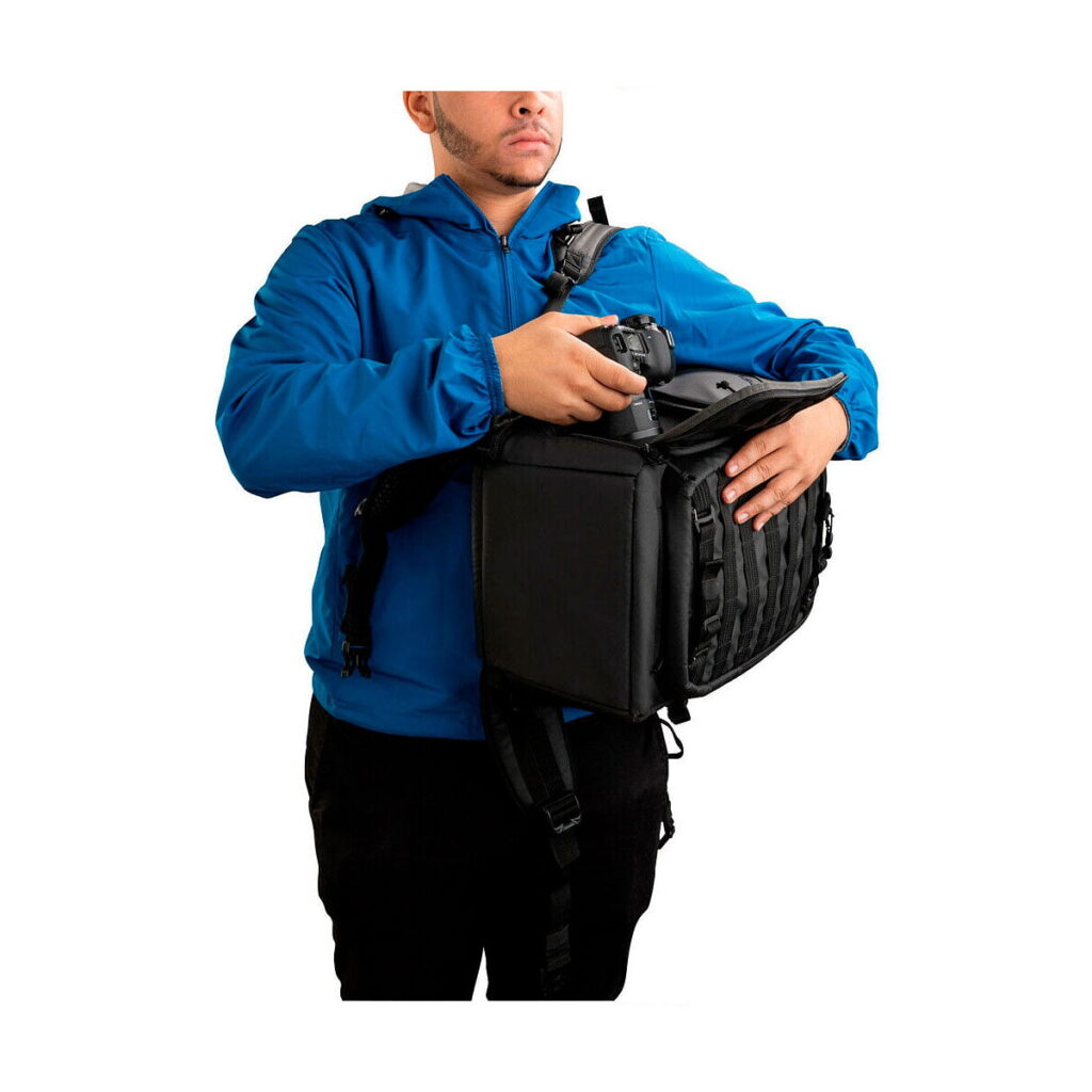 Tenba_Axis_20L_Tactical_Backpack_009 Siniseen takkiin pukeutunut henkilö hakee kameran Tenba Axis 20L Tactical Backpack -reppuunsa, jota hän kantaa edessään.