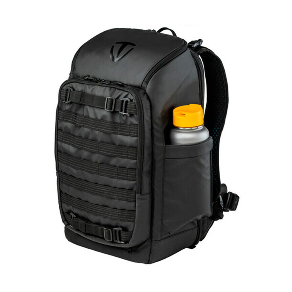 Tenba_Axis_24L_Tactical_Backpack_008 Tenba Axis 24L Tactical Backpack, musta, jossa on useita lokeroita ja etuhihnat, näkyy valkoista taustaa vasten, ja sen sivutaskussa on keltaisella korkilla varustettu vesipullo.