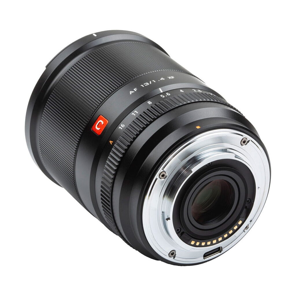 Viltrox_AF_13mm_f_1_4_Fuji_X_004 Lähikuva Viltrox AF 13mm f/1.4 -objektiivi, Fuji X näyttää sen mustan linssin, metallisen kiinnikkeen, elektroniset koskettimet ja merkityt aukko- ja tarkennusrenkaat valkoista taustaa vasten.