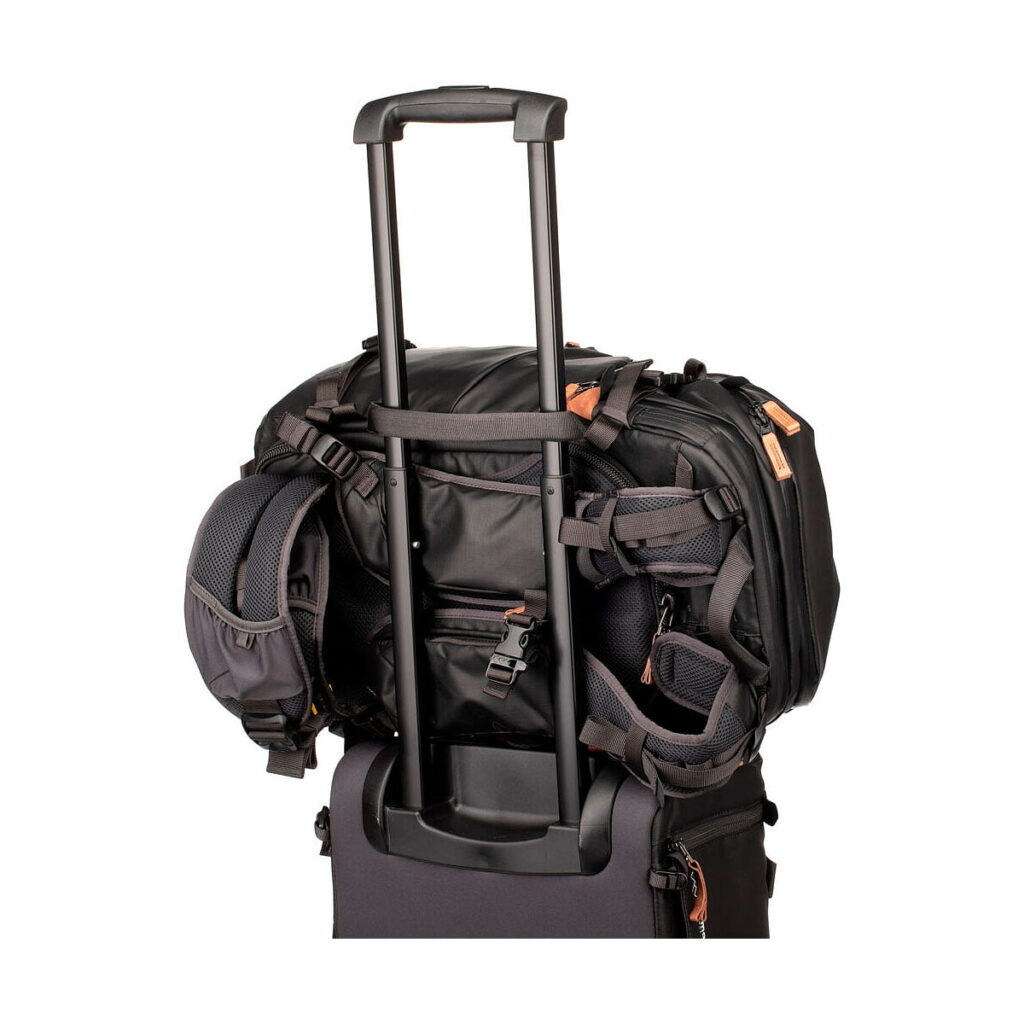 Shimoda Designs Explore v2 25 Starter Kit -kamerareppu, musta on kiinnitetty turvallisesti rullalaukun kahvaan, kuvassa takaa valkoista taustaa vasten. Mustassa repussa näkyy useita taskuja ja hihnoja.