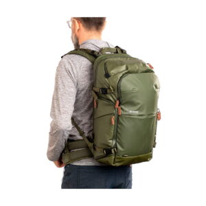 Henkilö, jolla on vaaleanharmaa pitkähihainen paita ja silmälasit, seisoo profiilissa ja kantaa Shimoda Designs Explore v2 30 -kamerareppua, vihreä - suurta oliivinvihreää reppua, jossa on ruskehtavat vetoketjut ja vyötäröhihna.