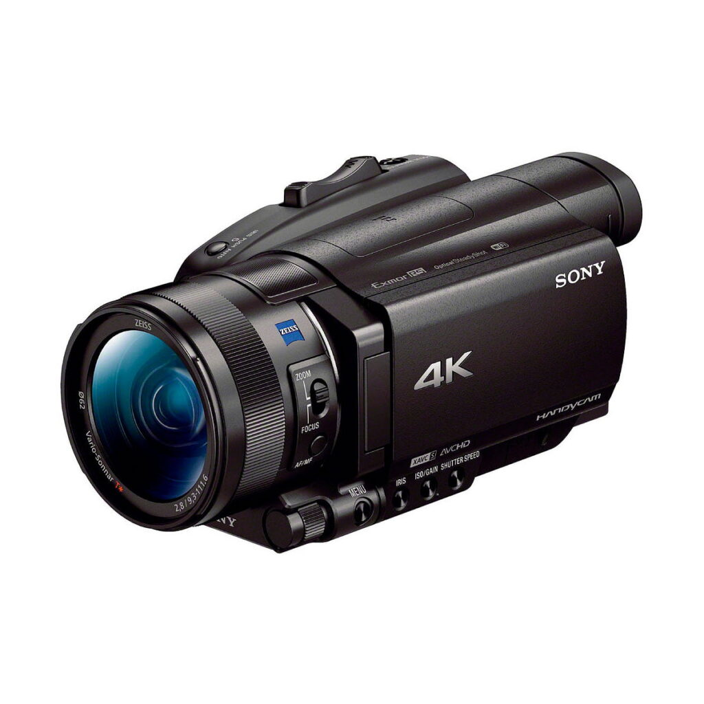 Sony_FDR-AX700_003 Musta Sony FDR-AX700 4K -videokamera, jossa on suuri objektiivi, useita ohjauspainikkeita ja käsikahva, näkyy etupuolelta valkoista taustaa vasten.