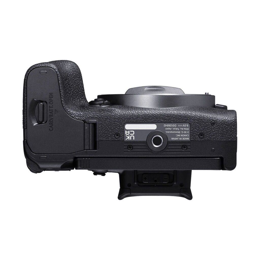 Canon_EOS_R10_004 Musta Canon EOS R10 -järjestelmäkamera alhaalta katsottuna, jossa näkyy akkulokero, jalustakiinnitys, tarrat, kuvioitu kahva ja osittain näkyvä objektiivi kuvan yläosassa.