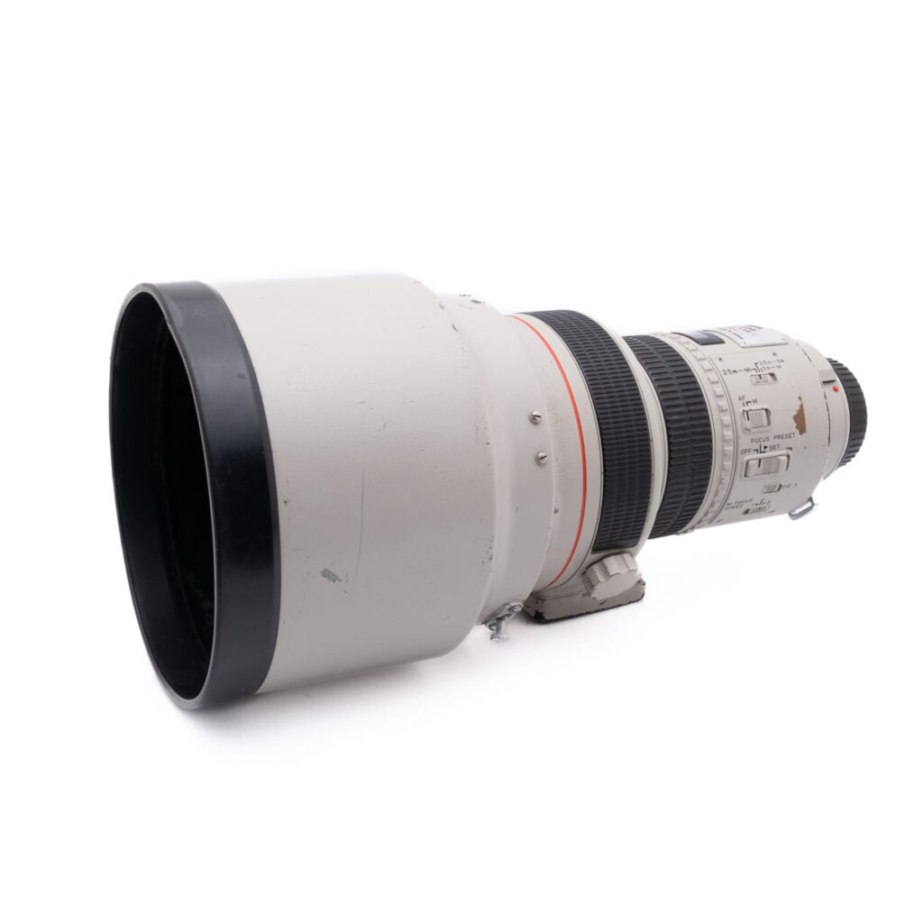 Canon 200mm f1.8L-1 Olympus OM-D E-M1X (SC 460) - Käytetty on esitetty valkoisella taustalla, ja siinä on suuri valkoinen teleobjektiivi, jossa on musta etureuna, yksityiskohtaiset tarkennusrenkaat ja zoomausrenkaat sekä useita säätökytkimiä.