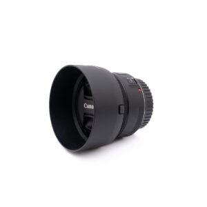 Musta Canon EOS M50 - Käytetty kamera, jossa on vastavalosuoja, makaa kyljellään valkoisella taustalla. Etuelementissä näkyy tuotemerkki Canon.