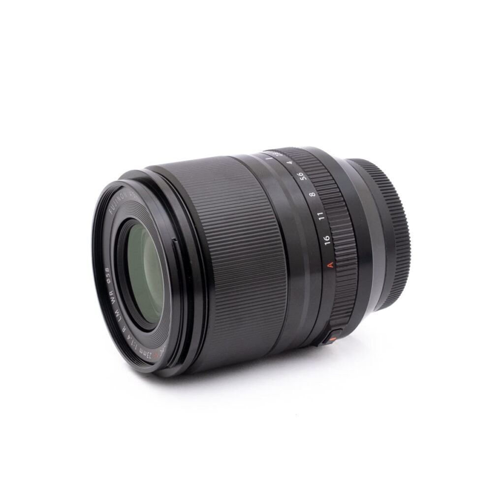 Fujinon 23mm f1.4-2 Olympus OM-D E-M1X (SC 460) - Käytetty kameran objektiivi, jossa on näkyvissä tarkennusrengas ja aukkorenkaat, lepää valkoisella taustalla, ja se on kulmailtu siten, että etulasi ja osa objektiivin piipusta tulevat näkyviin.