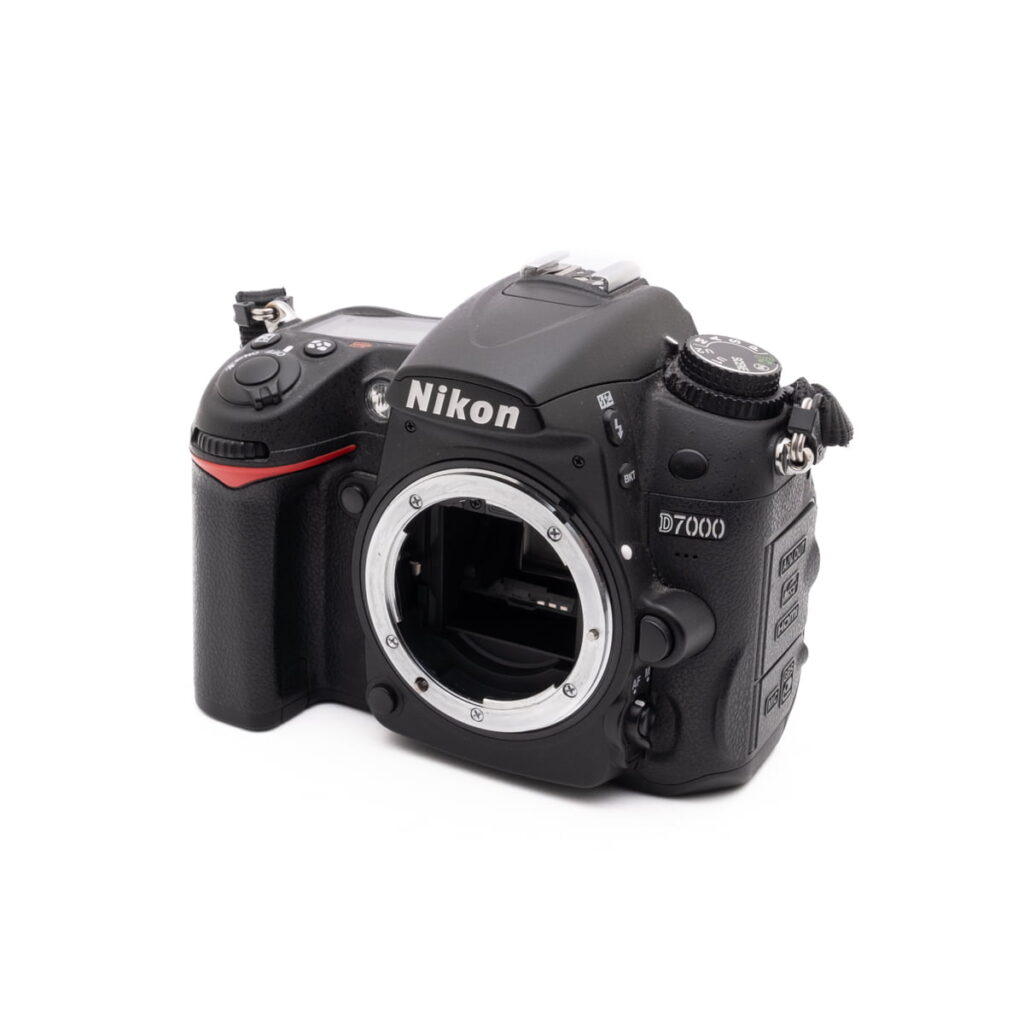 Nikon D7000-2 Musta Nikon D7000 (SC 7000) - Käytetty DSLR-kameran runko näkyy valkoisella taustalla, ja sen objektiivin kiinnitys on näkyvissä, ja sen sivussa ja yläreunassa on erilaisia ohjauspainikkeita ja säätimiä.