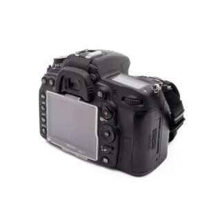 Nikon D7000-3 Nikon D7000 (SC 7000) - Käytetty digitaalinen DSLR-kamera, musta, takaa katsottuna, näyttö, painikkeet ja kädensija sekä hihna näkyvät valkoista taustaa vasten.