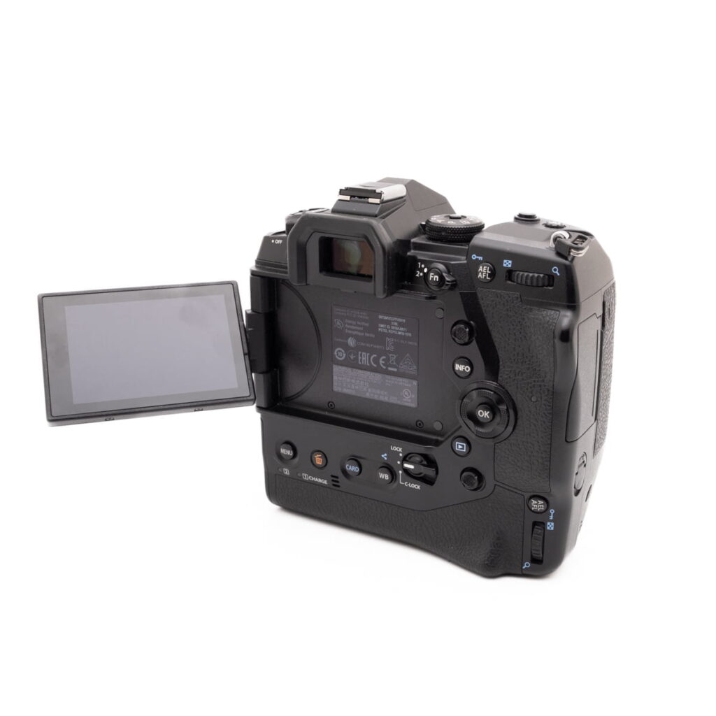 Olympus OM-D E-M1X-3 Olympus OM-D E-M1X (SC 460) - Käytetty on kuvattu takaa, ja siinä on kuvioitu musta runko, useita ohjauspainikkeita ja kallistettava LCD-näyttö vasemmalle.