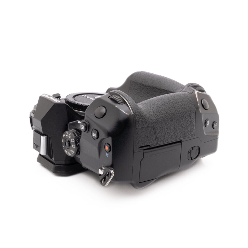 Olympus OM-D E-M1X-4 Olympus OM-D E-M1X (SC 460) - Käytetty -kameran ylhäältä kuvattu kuvakulma, jossa näkyy mustat kuvioidut kädensijat, ohjauspainikkeet, valitsimet ja osa objektiivin kiinnityksestä valkoista taustaa vasten.