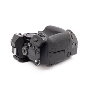 Olympus OM-D E-M1X-4 Olympus OM-D E-M1X (SC 460) - Käytetty -kameran ylhäältä kuvattu kuvakulma, jossa näkyy mustat kuvioidut kädensijat, ohjauspainikkeet, valitsimet ja osa objektiivin kiinnityksestä valkoista taustaa vasten.