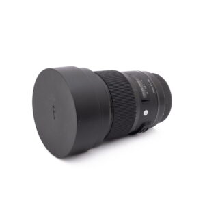 Sigma 20mm f1.4 Canon-2 Olympus OM-D E-M1X (SC 460) - Käytetty kamera on esitetty vaakasuorassa mustalla objektiivilla ja vastavalosuojalla valkoisella pohjalla.