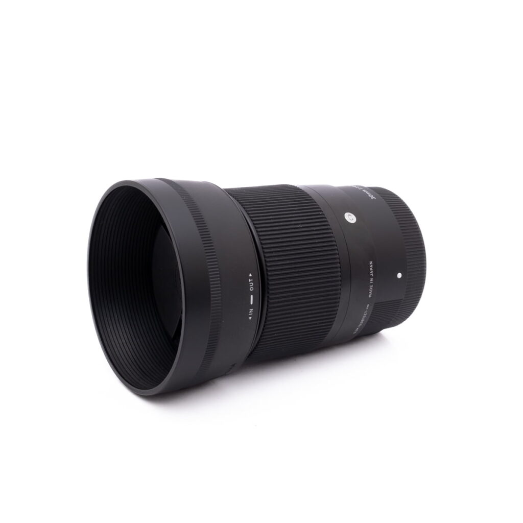 Sigma 30mm f1.4 SonyE-1 Sigma 30mm f/1.4 DC DN Sony E - Käytetty musta kameraobjektiivi, jossa on uurrettu tarkennusrengas ja vastavalosuoja, on kuvattu valkoisella taustalla hieman vasemmalle suunnattuna.