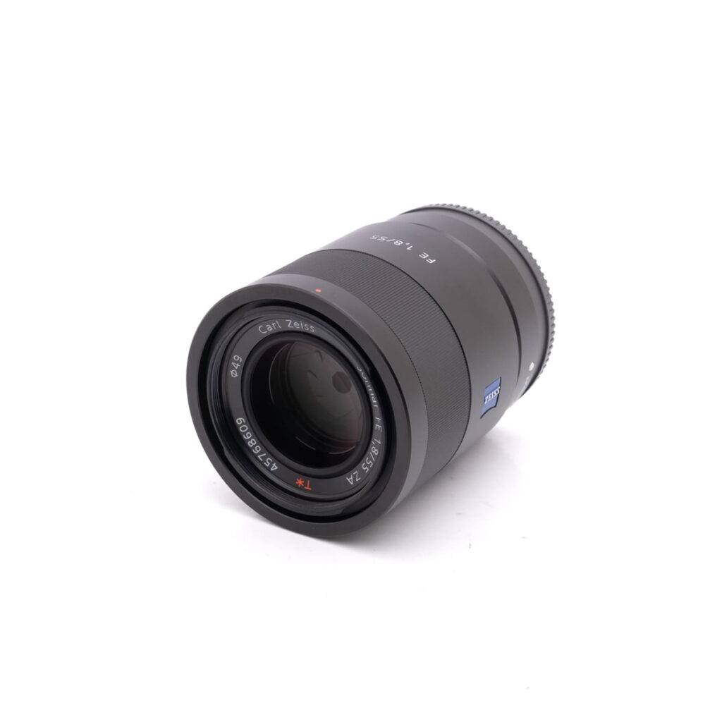 Sony-55mm-f1.8-2 Musta Sony FE Sonnar T* 55mm f/1.8 ZA - Käytetty -objektiivi on suunnattu valkoiselle taustalle, ja siinä näkyy etulasin elementti ja tuotemerkit.