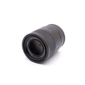 Sony-55mm-f1.8-2 Musta Sony FE Sonnar T* 55mm f/1.8 ZA - Käytetty -objektiivi on suunnattu valkoiselle taustalle, ja siinä näkyy etulasin elementti ja tuotemerkit.