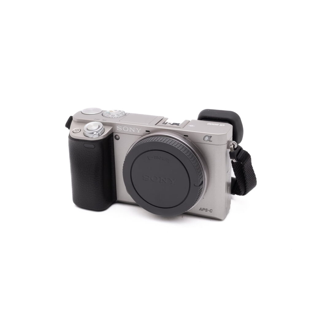 Sony-A6000-1 Hopea Sony a6000 (SC 470, K4.5) - Käytetty APS-C peilitön kamera, jossa on musta kahva, objektiivin kiinnityssuoja ja musta hihna kiinnitettynä, valkoisella taustalla.