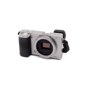 Sony-A6000-2 Hopeinen Sony a6000 (SC 470, K4.5) - Käytetty APS-C-peilittömän kameran runko ilman objektiivia, jossa on valotettu kuvakenno, musta kahva vasemmalla ja musta hihna oikealla.