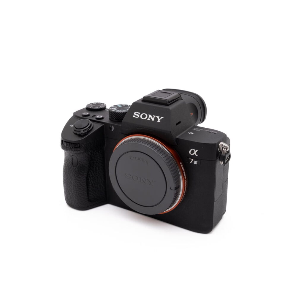 Sony-A7III-274-1 Käytetty Sony A7 III (SC 2800, Kunto K5) peilitön digitaalikamerarunko, musta ja objektiivin suojus, vinossa valkoisella taustalla.