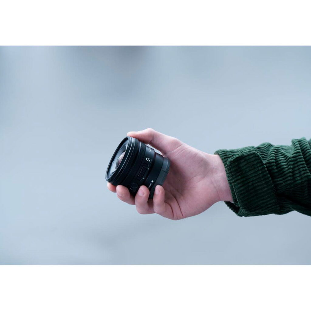 Sony-E-PZ-10-20mm-f4-G-4 Vihreään takkiin pukeutunut henkilö pitää Sony E PZ 10-20mm f/4 G -objektiivia oikeassa kädessään vaaleansinistä taustaa vasten.