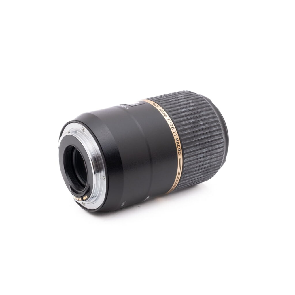 Tamron SP 90mm f/2.8 Macro Canon EF - Käytetty -objektiivi, jossa on kuvioitu tarkennusrengas ja kultaiset korostukset, makaa kyljellään, jolloin objektiivin kiinnitys ja elektroniset koskettimet näkyvät valkoista taustaa vasten.