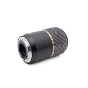 Tamron SP 90mm f/2.8 Macro Canon EF - Käytetty -objektiivi, jossa on kuvioitu tarkennusrengas ja kultaiset korostukset, makaa kyljellään, jolloin objektiivin kiinnitys ja elektroniset koskettimet näkyvät valkoista taustaa vasten.