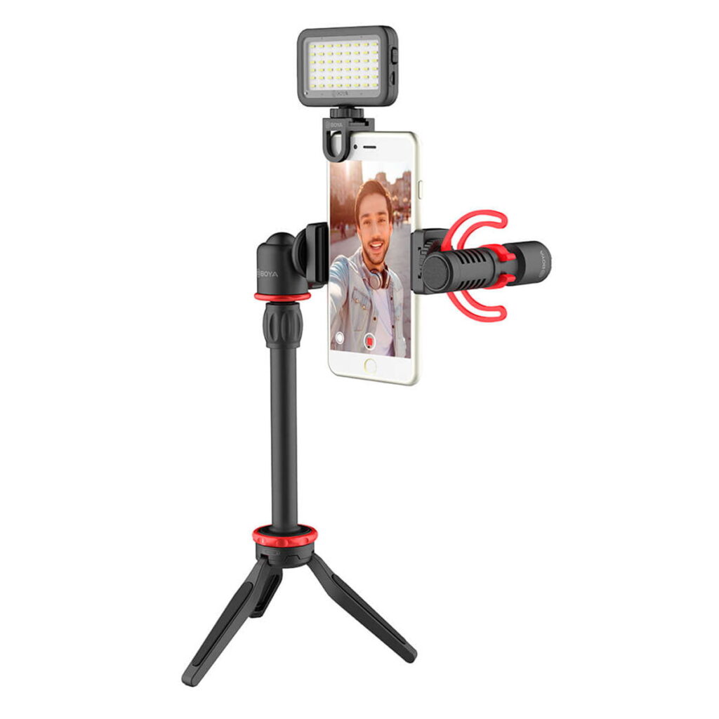 Boya-BY-VG350-3 Boya BY-VG350 Video-Kit pitää älypuhelinta mustapunaisella jalustalla, johon on kiinnitetty LED-valo ja mikrofoni vloggausta tai videokuvausta varten. Puhelimen näytöllä näkyy selfietä ottava mies.
