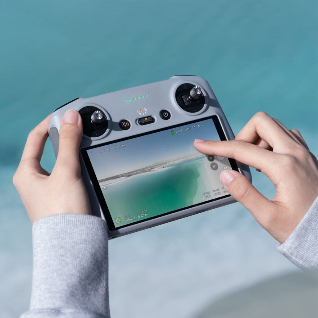 Henkilö käyttää DJI RC Smart Controller -ohjainta -ohjain, jonka sisäänrakennettu näyttö näyttää ilmasta ranta- ja merinäkymän, todennäköisesti ohjaamassa lennokkia lähellä rantaviivaa.