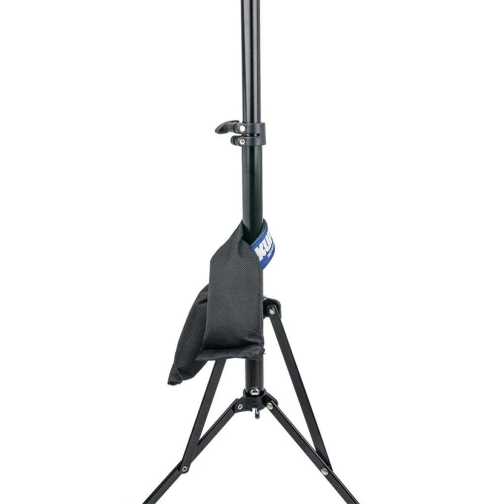 Kupo-KSW-05-4 Musta kolmijalkainen valaisinjalusta, jonka korkeus on säädettävissä ja jossa on vakauden vuoksi toiseen jalkaan kiinnitetty Kupo KSF-15 Shot Bag (6,9 kg). Asetelma on eristetty valkoiselle taustalle.