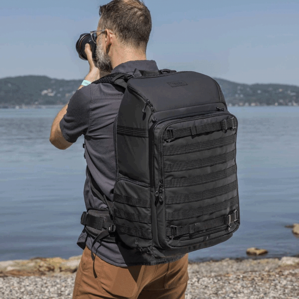 Tenba-Axis-V2-32L-5 A bearded man stands on a rocky shore facing the lake and photographs the view, wearing a Tenba Axis V2 32L backpack. Across the water, forested hills under a clear sky.