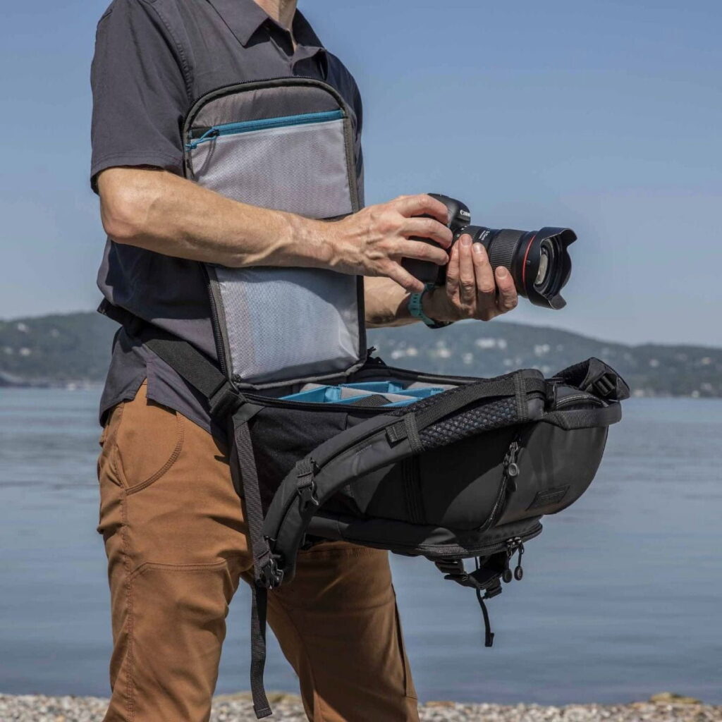 Tenba-axis V2-16L-2 A person stands on the water holding a camera, wearing a Tenba Axis V2 16L Backpack with an open front flap revealing its organised interior space for camera gear. The backpack is secured for easy access.