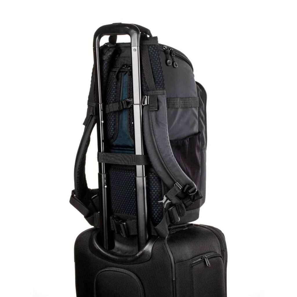 Tenba-axis V2-16L-6 The Tenba Axis V2 16L camera backpack is securely attached to the handle of the black roller bag for easy transport. The background is plain white.