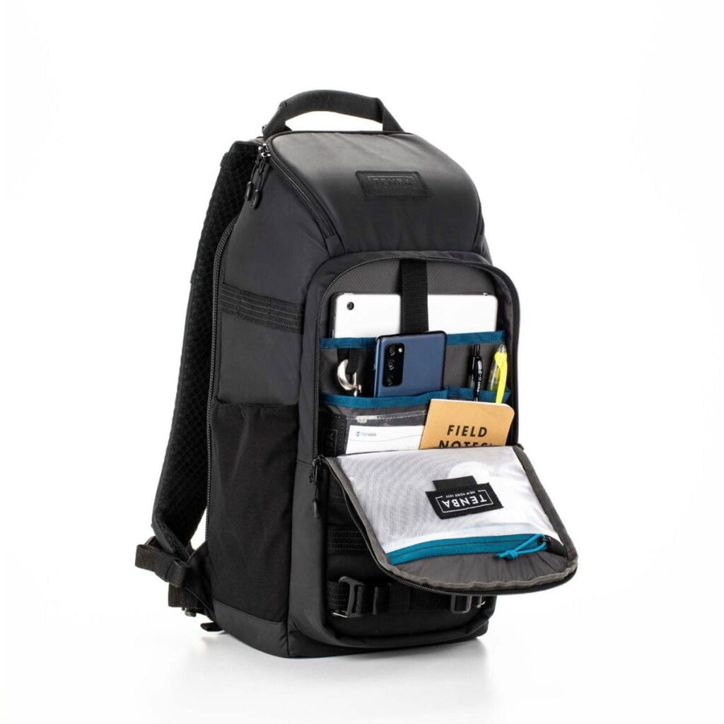 Tenba-axis V2-16L-8 The Tenba Axis V2 16L Backpack stands upright with the front pocket open with a zipper and displays organized compartments for a phone, tablet, pen, notebook and mesh pouch with TENBA labeling. It has padded straps and top handle.