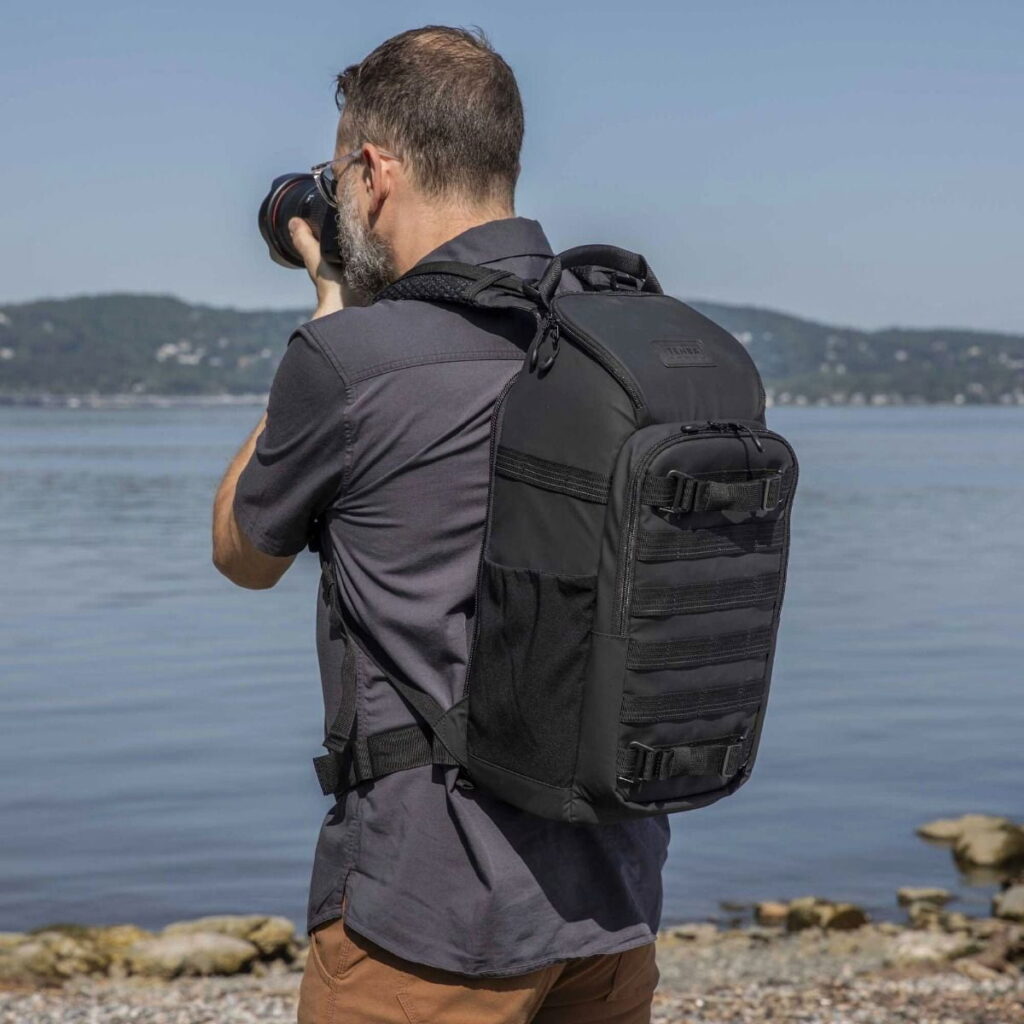 Tenba-axis V2-16L-9 A bearded man in a dark shirt and brown trousers stands on the shore of a lake with the hills in the background, taking photos and carrying a Tenba Axis V2 16L Backpack.